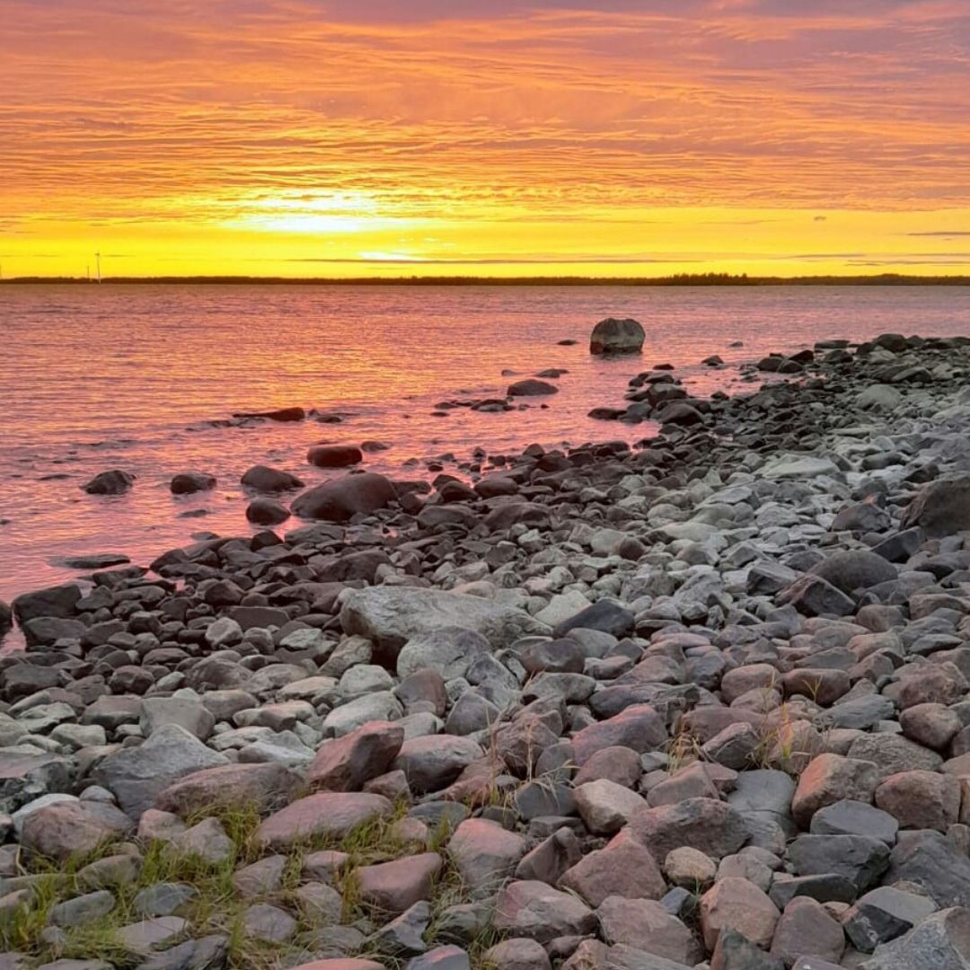Auringonlasku meren rannalla. Taivas värjäytyy oranssin keltaiseksi ja meren pinta violetiksi. Etualalla kivikkoinen ranta.